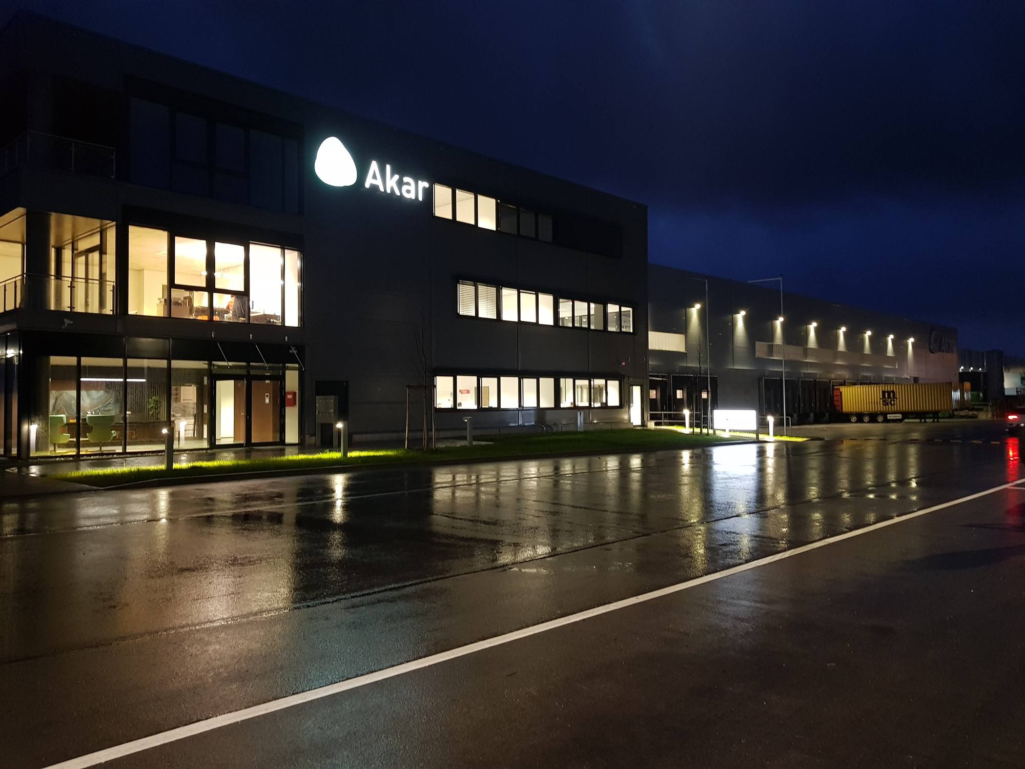 Dasselbe Gebäude zwei Tage später bei Nacht: Logo und Schriftzug „Akar“ leuchten weiß, nasse Straße spiegelt das Licht