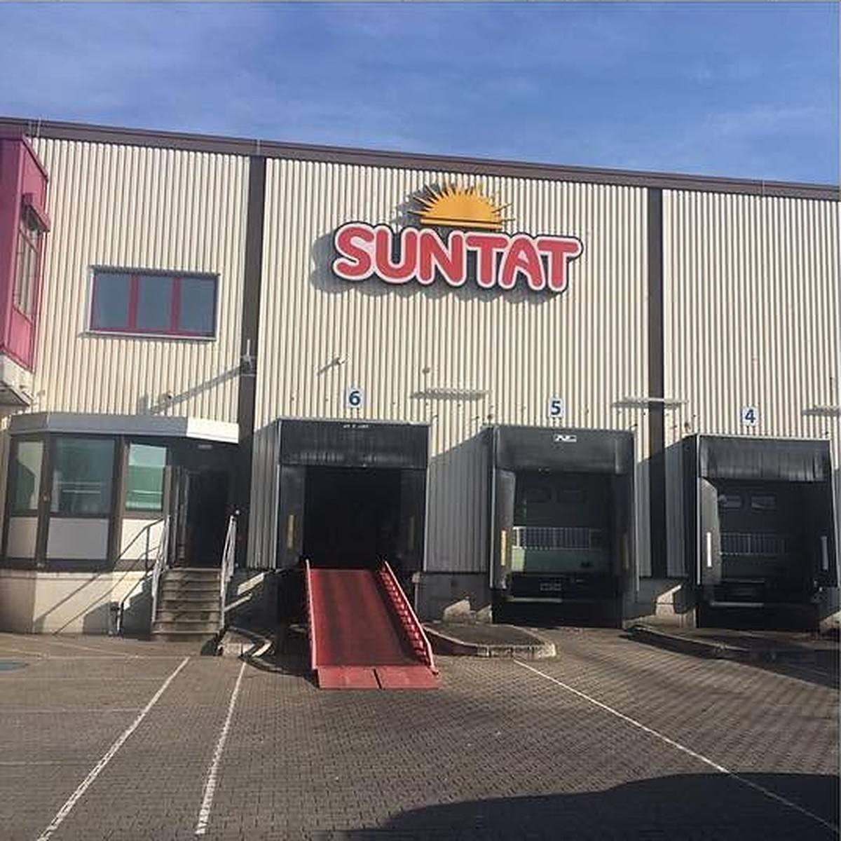 Lagerhalle mit Laderampen und Leuchtschrift „SUNTAT“ mit gelbem Sonnenlogo bei Tag