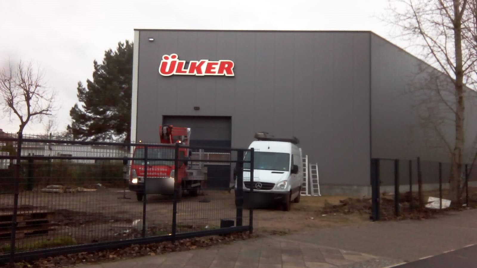 Graue Lagerhalle mit rotem Leuchtschriftzug „ÜLKER“, davor Hubarbeitsbühne und Transporter bei Tag